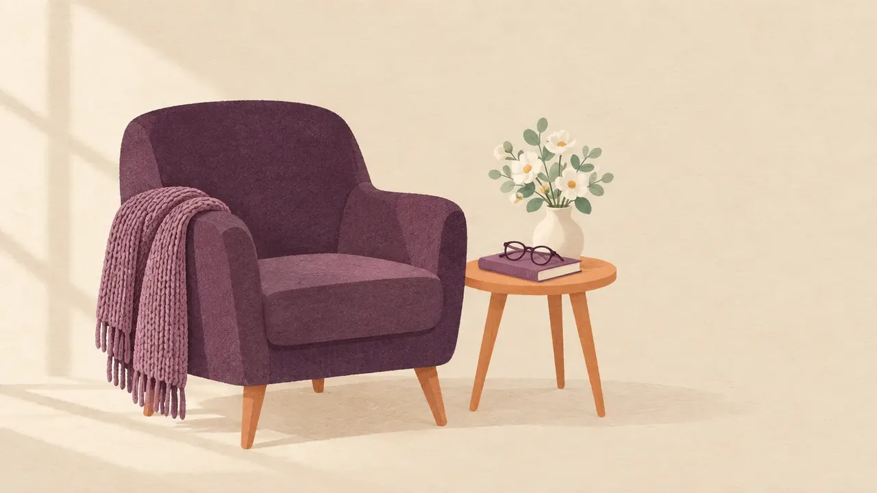 An empty armchair with a folded knitted blanket over one arm, reading glasses resting on a closed book and a small vase of white flowers.